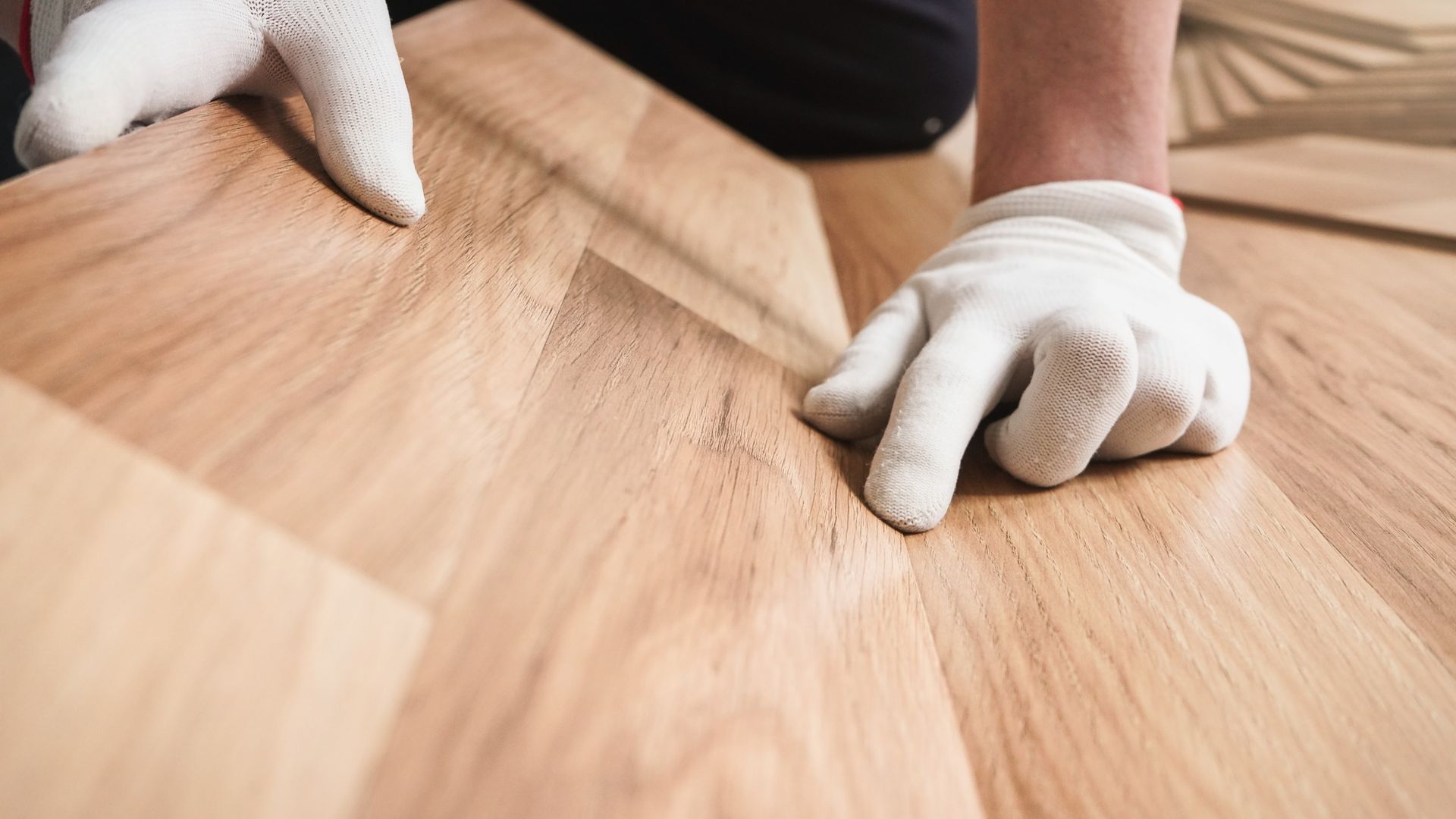 A person wearing white work gloves installs light-colored wooden floor planks. A person wearing white work gloves installs light-colored wooden floor planks.