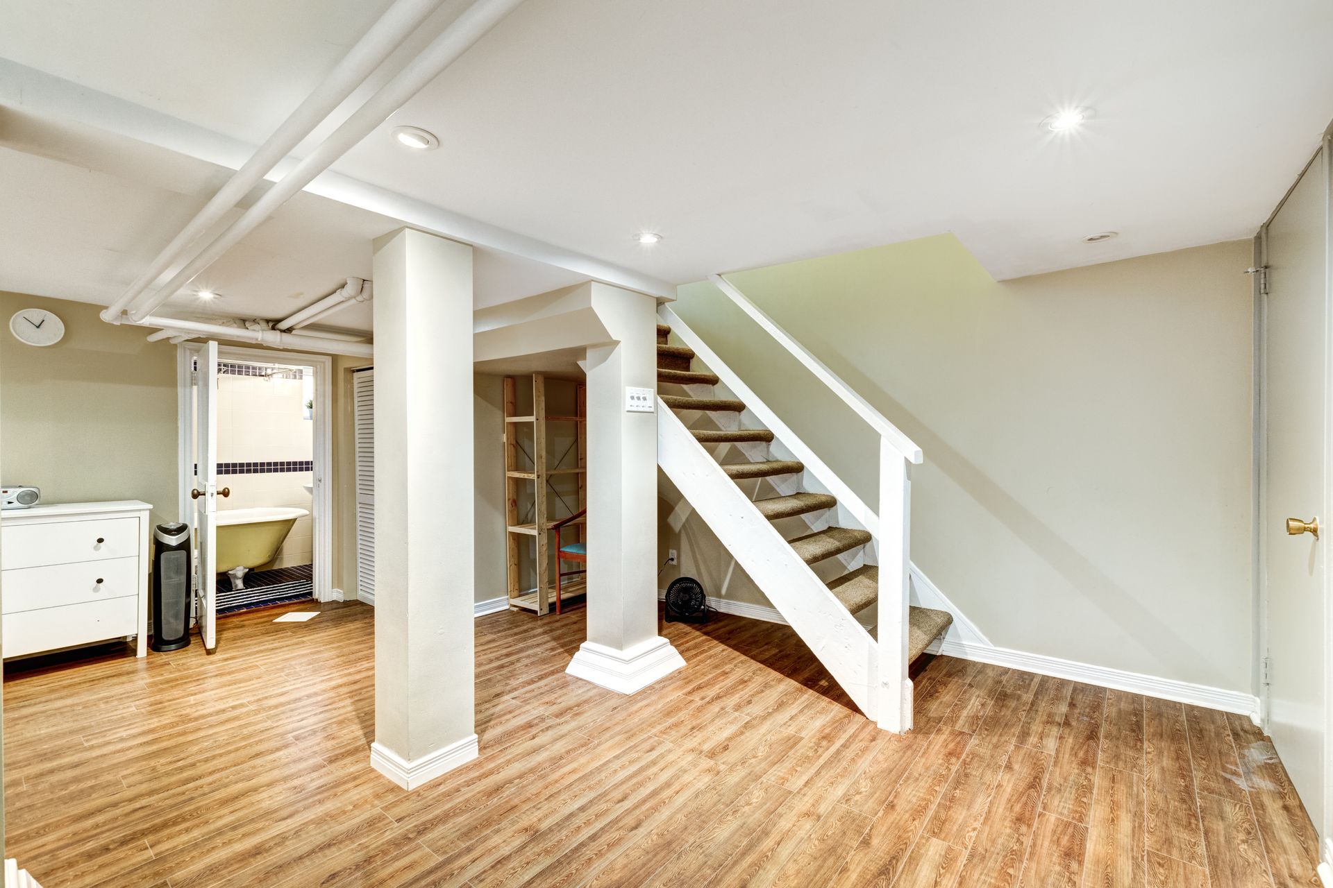 A finished basement with wood-look flooring, two central support columns, white stairs, and a doorway to a bathroom.