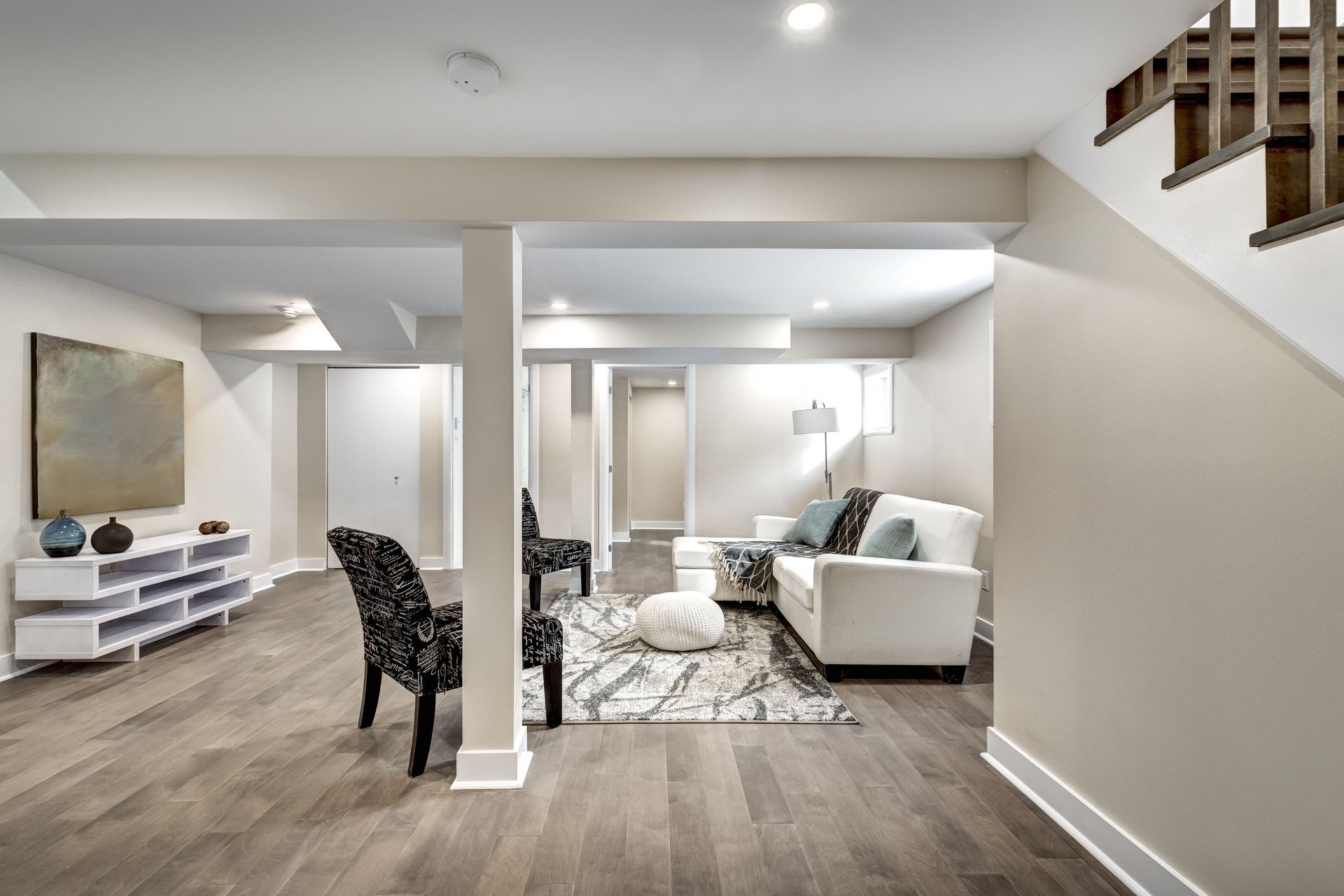 A finished basement with light wood floors, a white sofa, patterned armchairs, and a modern white media console. A finished basement with light wood floors, a white sofa, patterned armchairs, and a modern white media console.