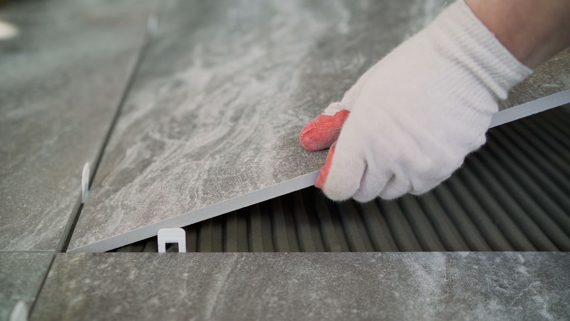A gloved hand places a gray marble-look tile onto a ridged layer of thin-set mortar, with tile leveling spacers visible. A gloved hand places a gray marble-look tile onto a ridged layer of thin-set mortar, with tile leveling spacers visible.