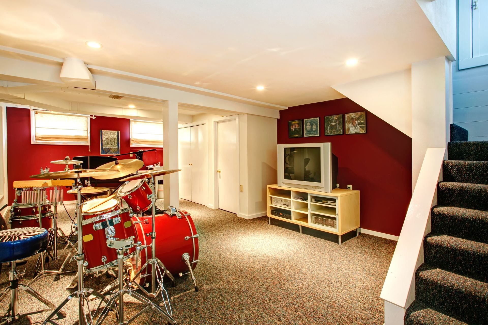 A drum kit in a basement with dark red walls, a television on a light wood stand, and stairs on the right.