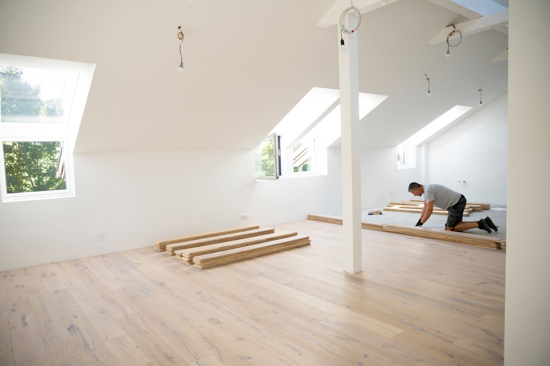 A worker kneels on a light-colored wooden floor in an unfinished attic, installing materials near skylights. A worker kneels on a light-colored wooden floor in an unfinished attic, installing materials near skylights.