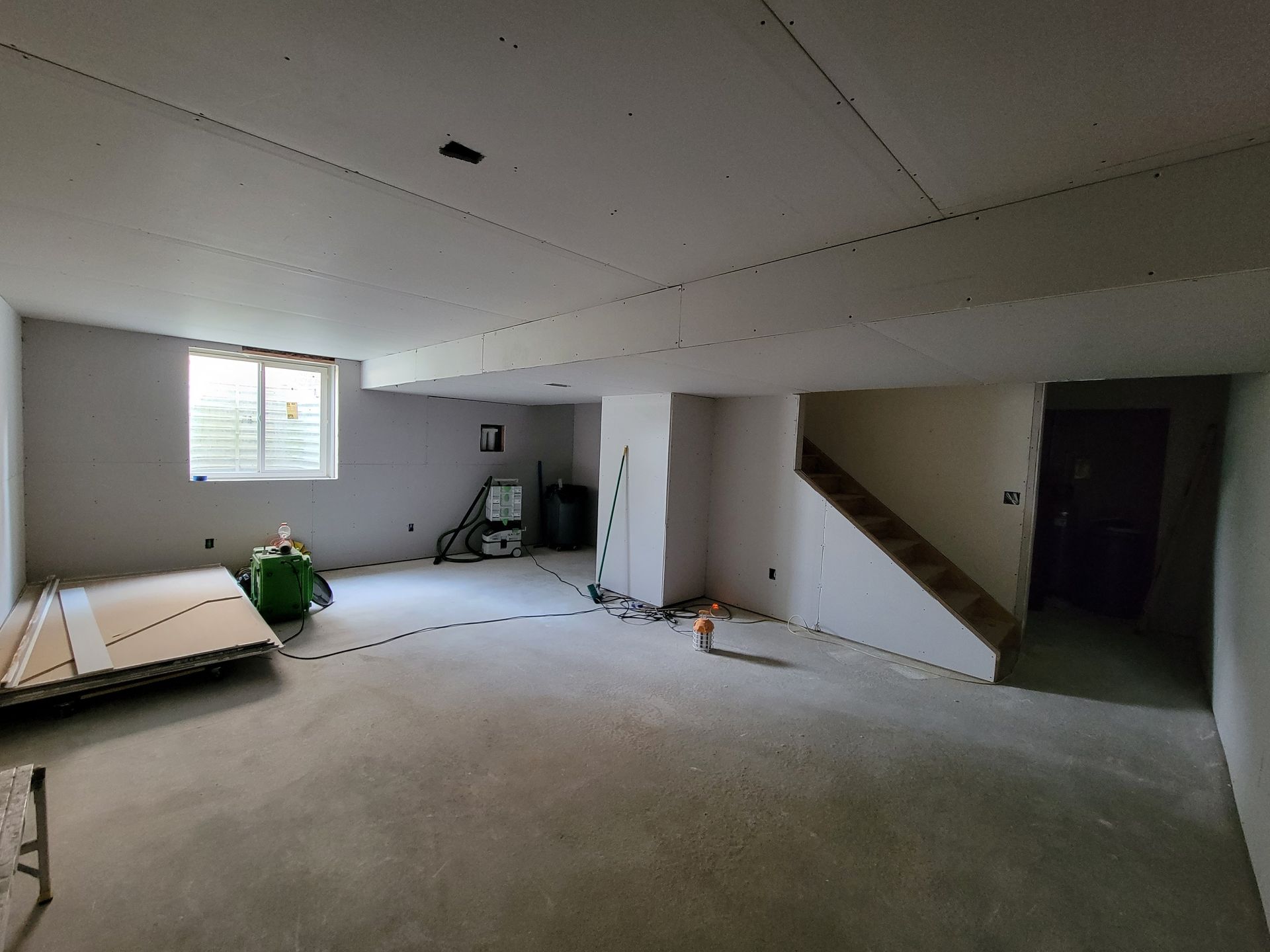 Unfinished basement with drywall, a small window, a staircase, and construction materials on a concrete floor. Unfinished basement with drywall, a small window, a staircase, and construction materials on a concrete floor.