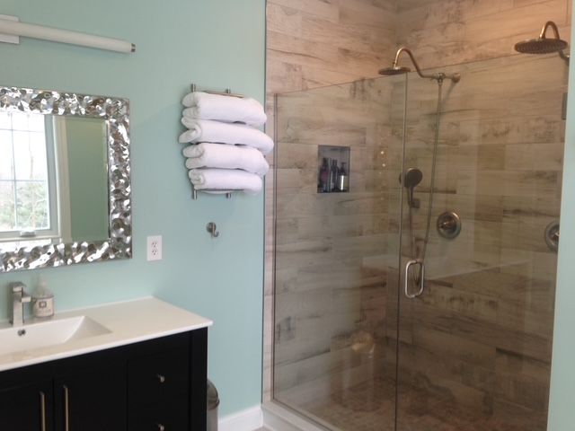 A bathroom with a black vanity, silver-framed mirror, stacked towels, and a walk-in shower with wood-look tile walls.
