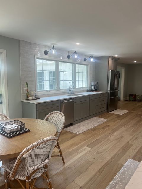 A modern kitchen with grey cabinets, light wood flooring, a central window, and a wooden dining table with woven chairs. A modern kitchen with grey cabinets, light wood flooring, a central window, and a wooden dining table with woven chairs.