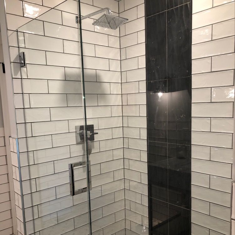A modern shower with white subway tiles, a vertical stripe of black accent tiles, a glass door, and a square showerhead. A modern shower with white subway tiles, a vertical stripe of black accent tiles, a glass door, and a square showerhead.