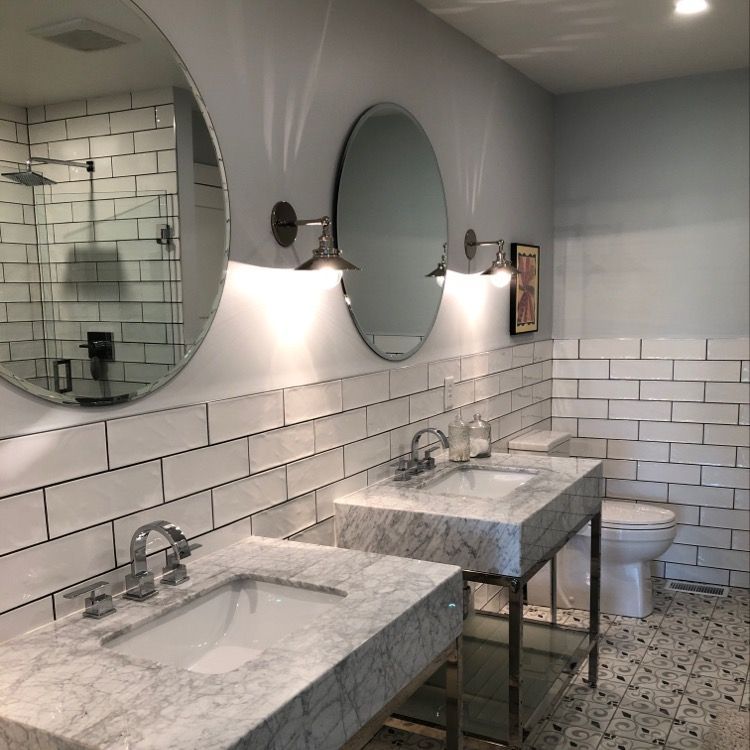 A modern bathroom with two marble pedestal sinks, oval mirrors, wall-mounted lights, and white subway tile walls.