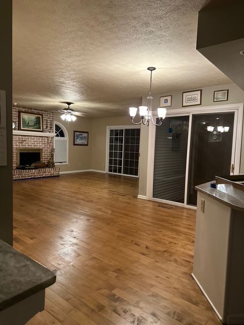 Spacious room with wood floors, a brick fireplace, sliding glass doors, and a chandelier, viewed from a kitchen counter.