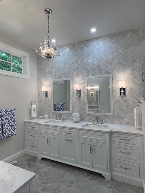 A double vanity bathroom with white cabinets, marble herringbone tile wall, two mirrors, sconces, and a crystal chandelier. A double vanity bathroom with white cabinets, marble herringbone tile wall, two mirrors, sconces, and a crystal chandelier.