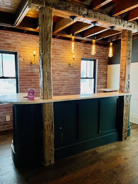 A dark green curved bar with a white countertop features rustic wood pillars and pendant lights against a brick wall.