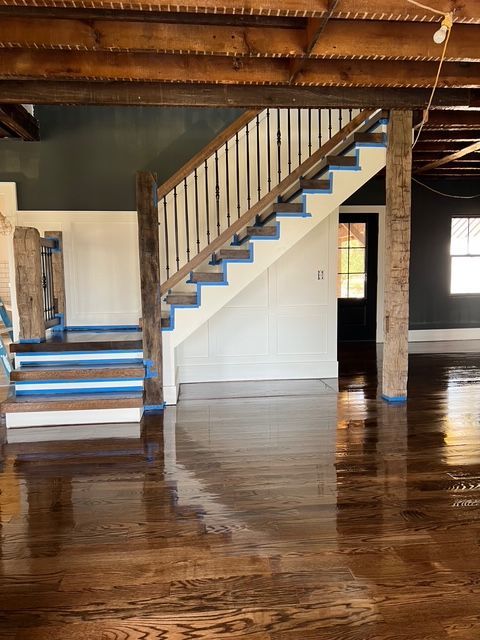 A staircase with painter's tape on the edges sits in an open room with dark hardwood floors and exposed wooden beams.