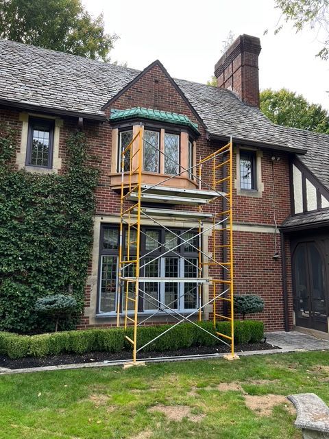 A brick Tudor-style house with a bay window features yellow scaffolding standing in front of its lower-level windows.