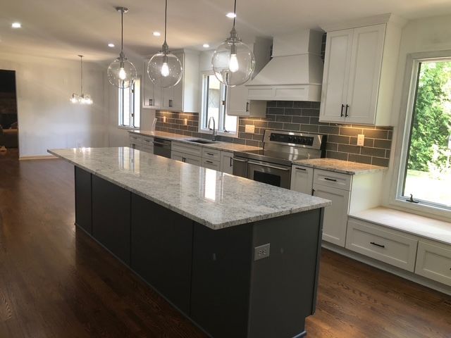 A modern kitchen featuring a dark gray island with white countertops, white cabinets, a gray tile backsplash, and pendants.