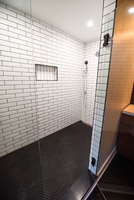 A walk-in shower with white subway tile walls, a small recessed niche, and a dark textured floor behind a glass door.