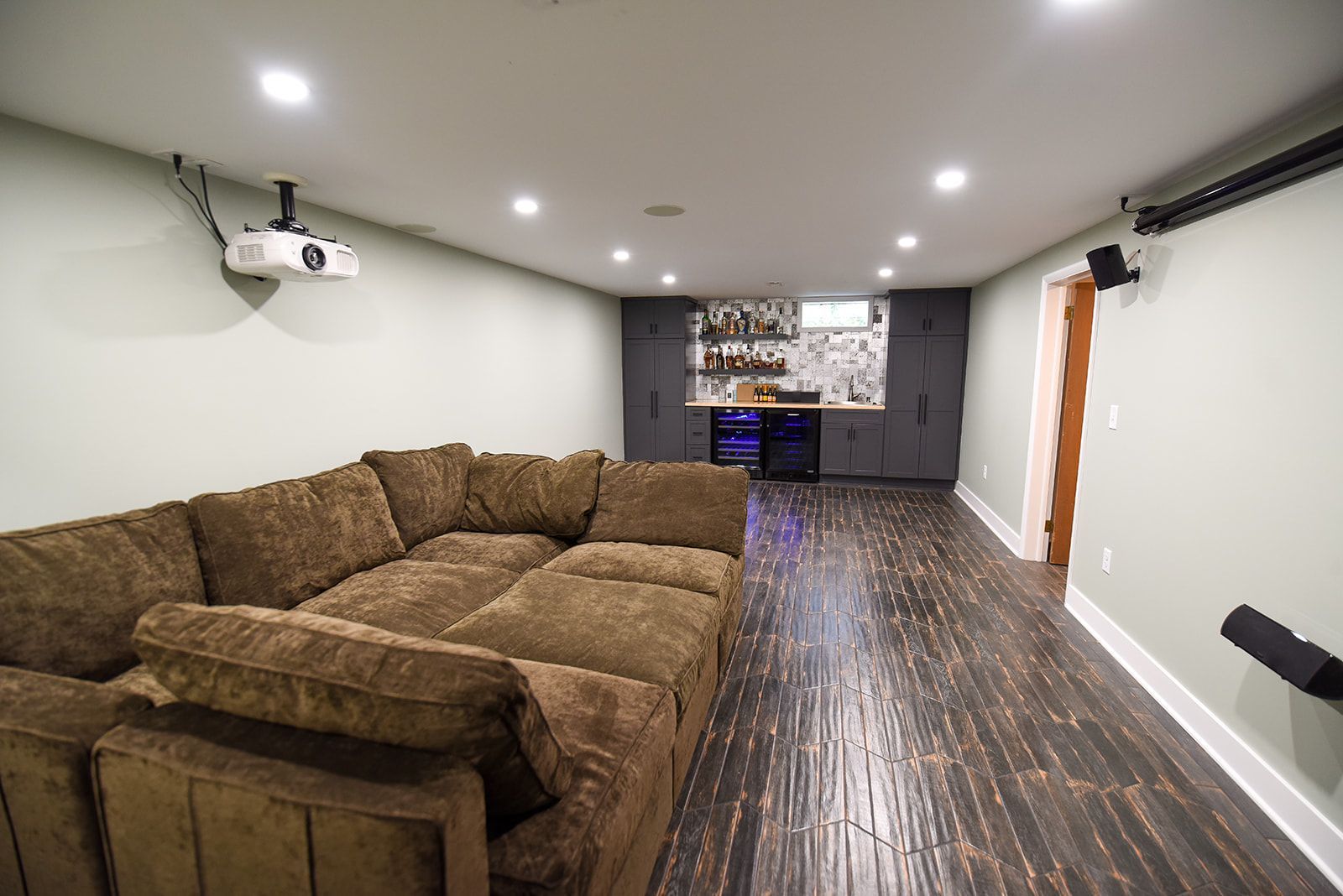 A home theater basement featuring a large brown sectional sofa, a projector, and a built-in bar area with dark cabinets.