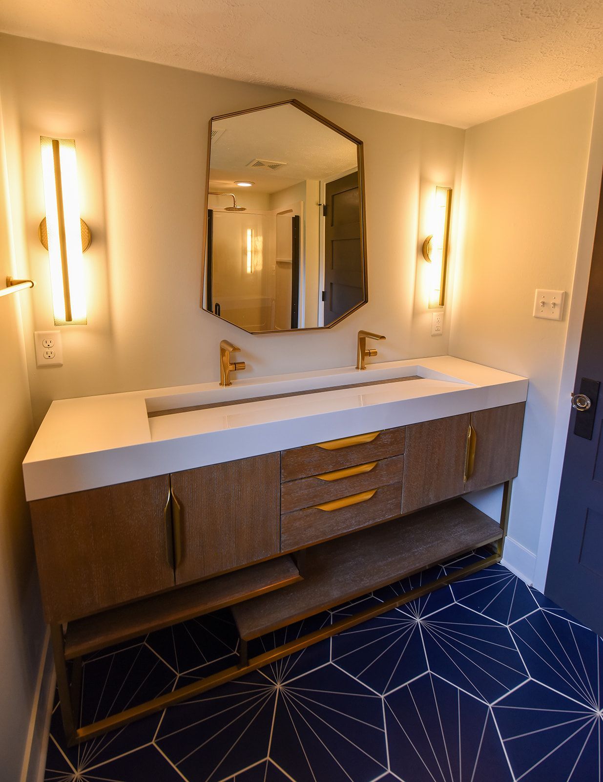 A modern wood vanity with a white double sink, gold fixtures, and a geometric mirror, set against dark geometric tile. A modern wood vanity with a white double sink, gold fixtures, and a geometric mirror, set against dark geometric tile.