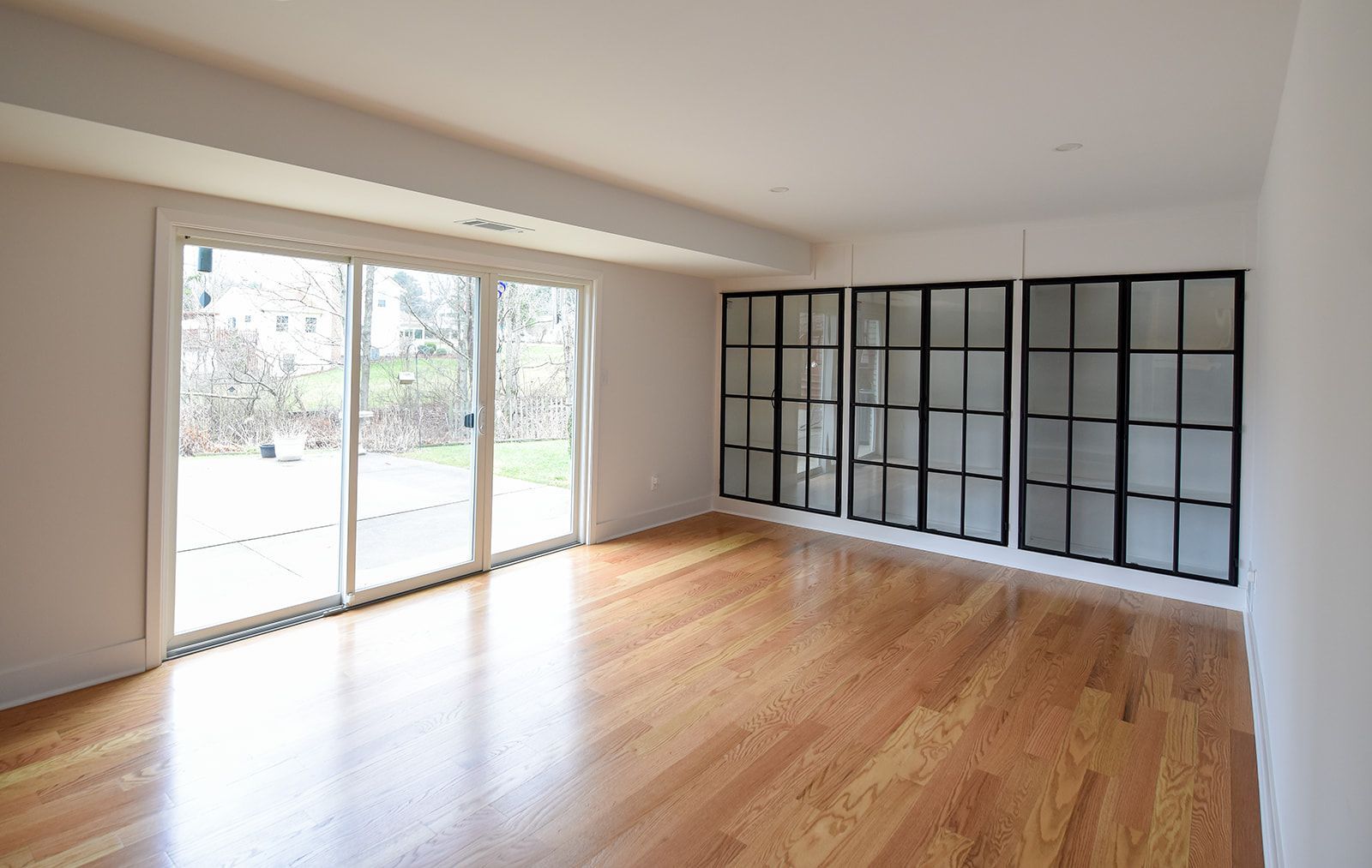 A sunlit room with hardwood floors, a large sliding glass door, and a wall of floor-to-ceiling windows with black frames.