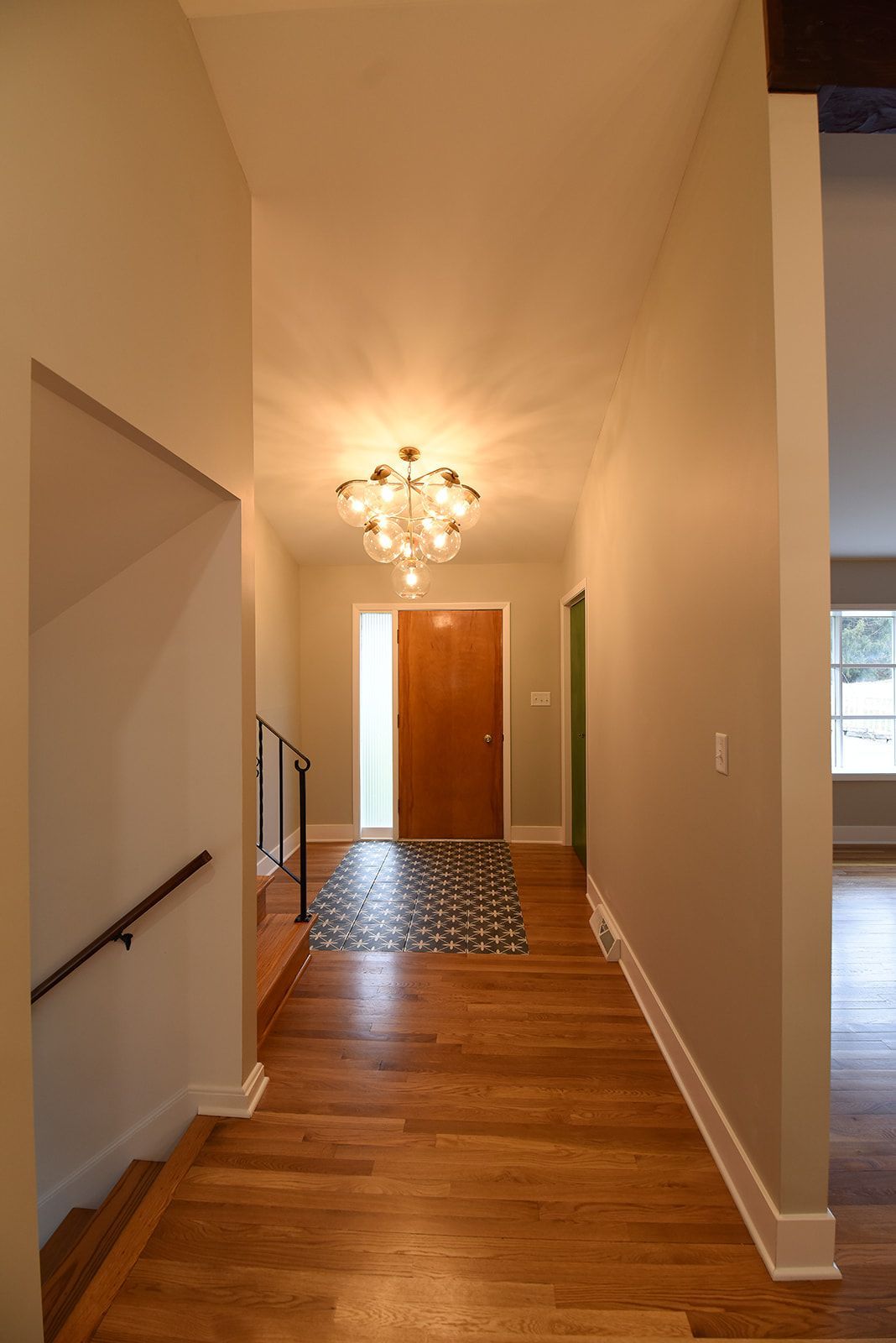 A hallway with hardwood floors leading to a front door with a decorative patterned tile floor under a modern chandelier.