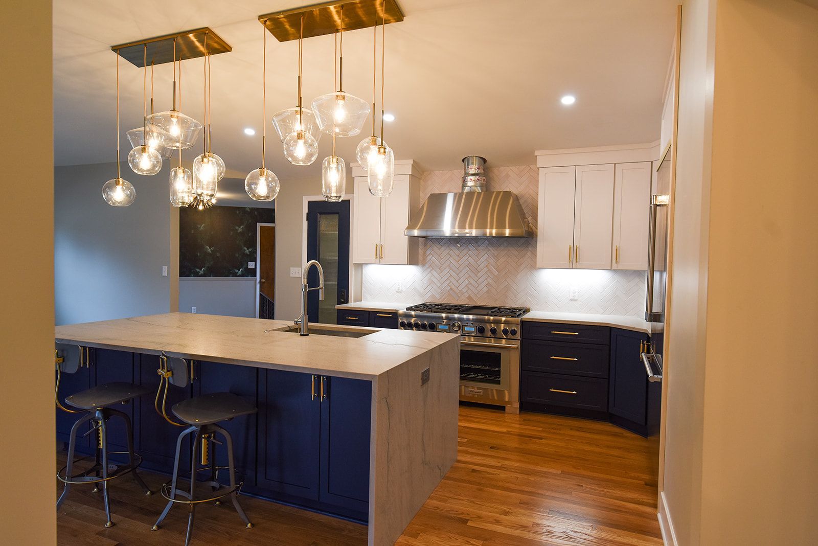 A kitchen featuring a blue island with bar stools, white upper cabinets, stainless steel appliances, and gold lighting.