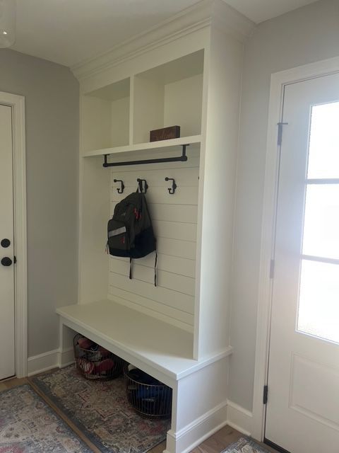 A white built-in entryway bench with shiplap backing, coat hooks, and overhead storage shelves between two doors.
