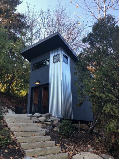 A small, modern cabin with dark gray walls and a corrugated metal panel, accessed by concrete steps in a wooded area.