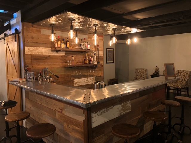 A rustic home bar with a metal countertop, wooden paneling, pendant lighting, and several bar stools. A rustic home bar with a metal countertop, wooden paneling, pendant lighting, and several bar stools.