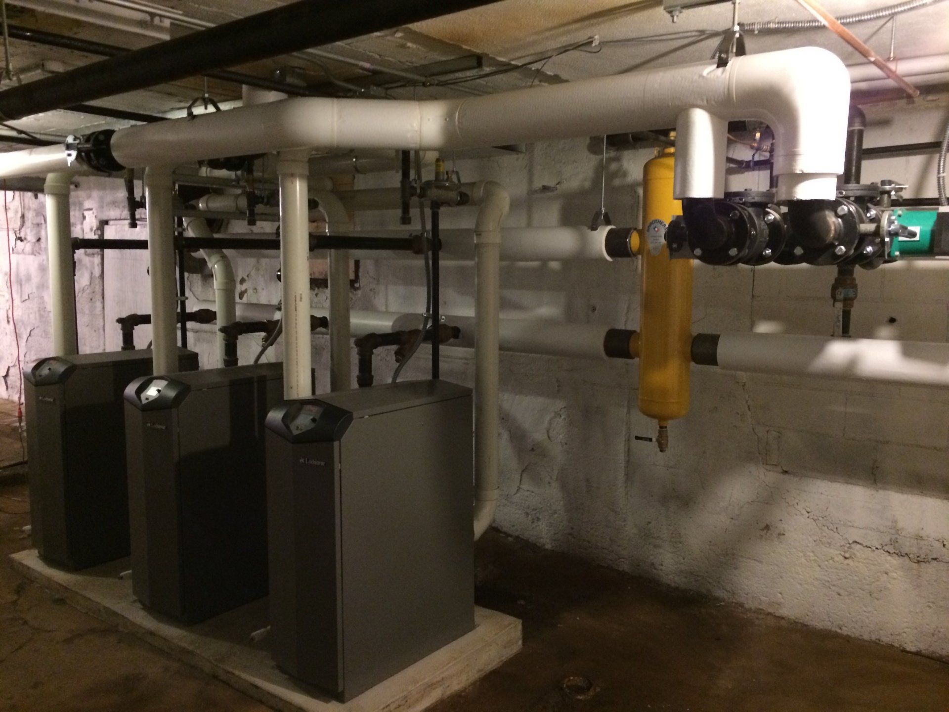 Three gray boilers with white pipes against a white brick wall in a basement.