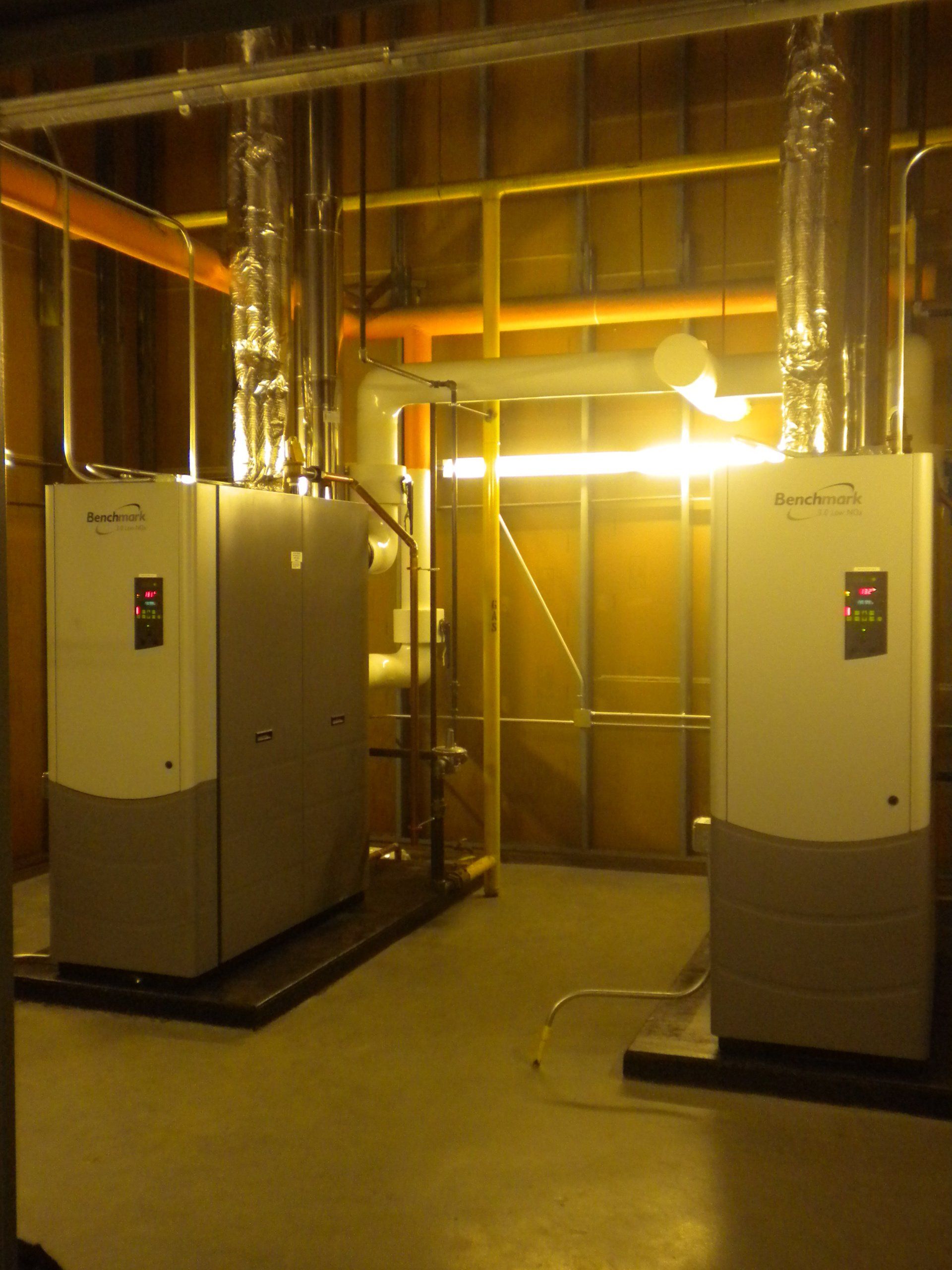 Two gray industrial heating units in a utility room, connected by pipes and venting up.