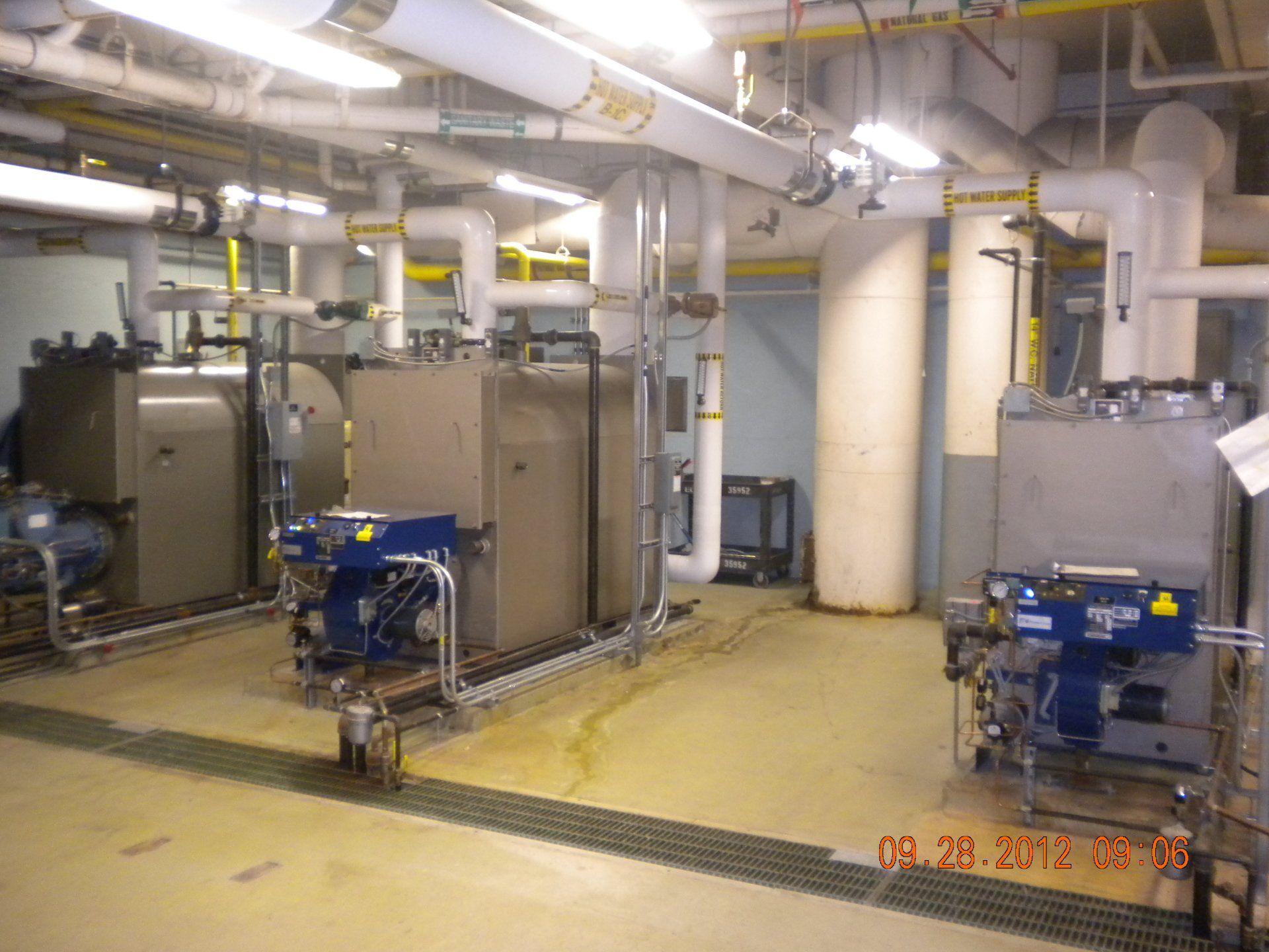 Industrial room with three boilers, pipes, and machinery. Gray and blue tones, indoors.