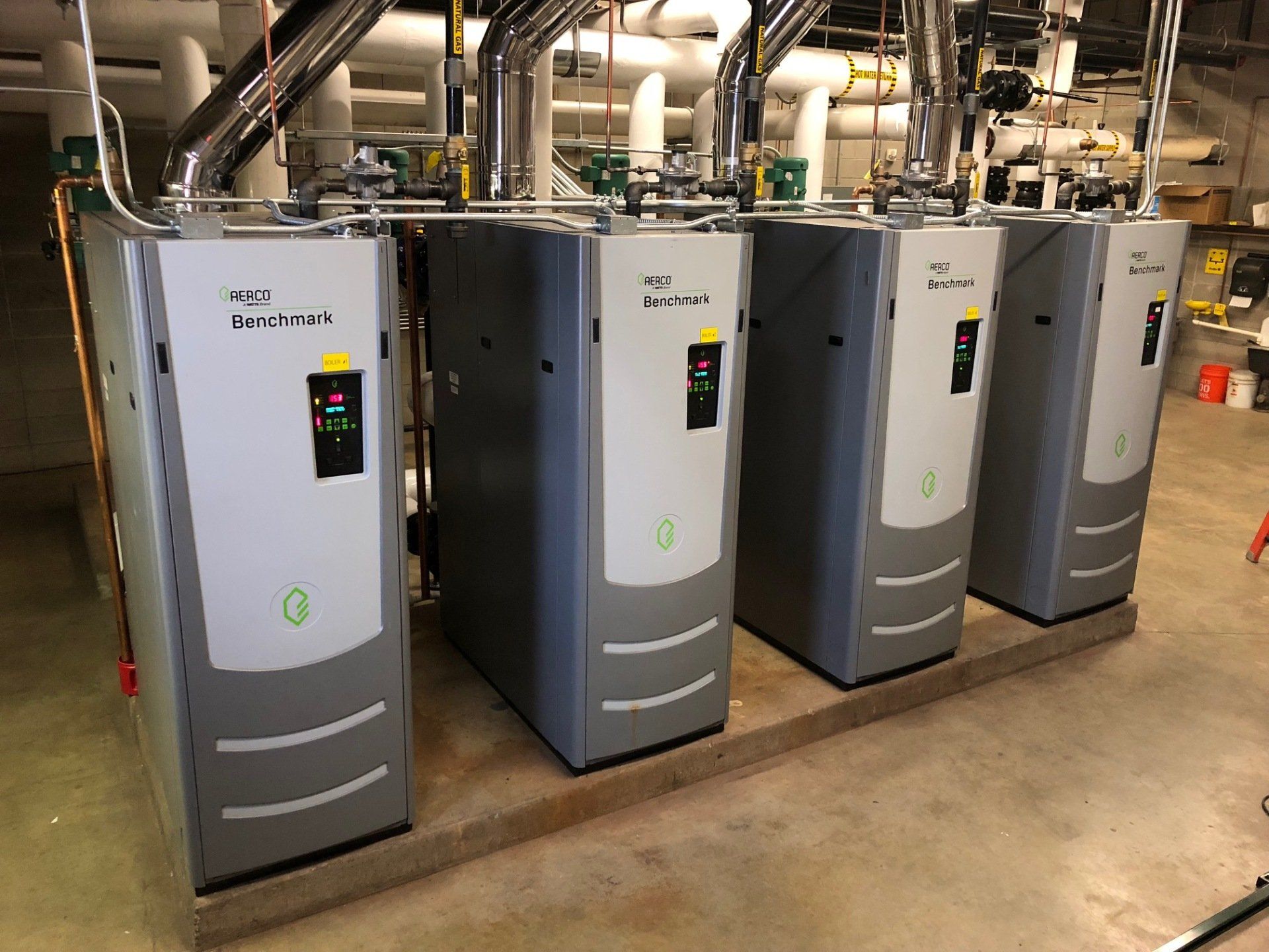 Four gray boilers in a utility room, connected to pipes overhead.