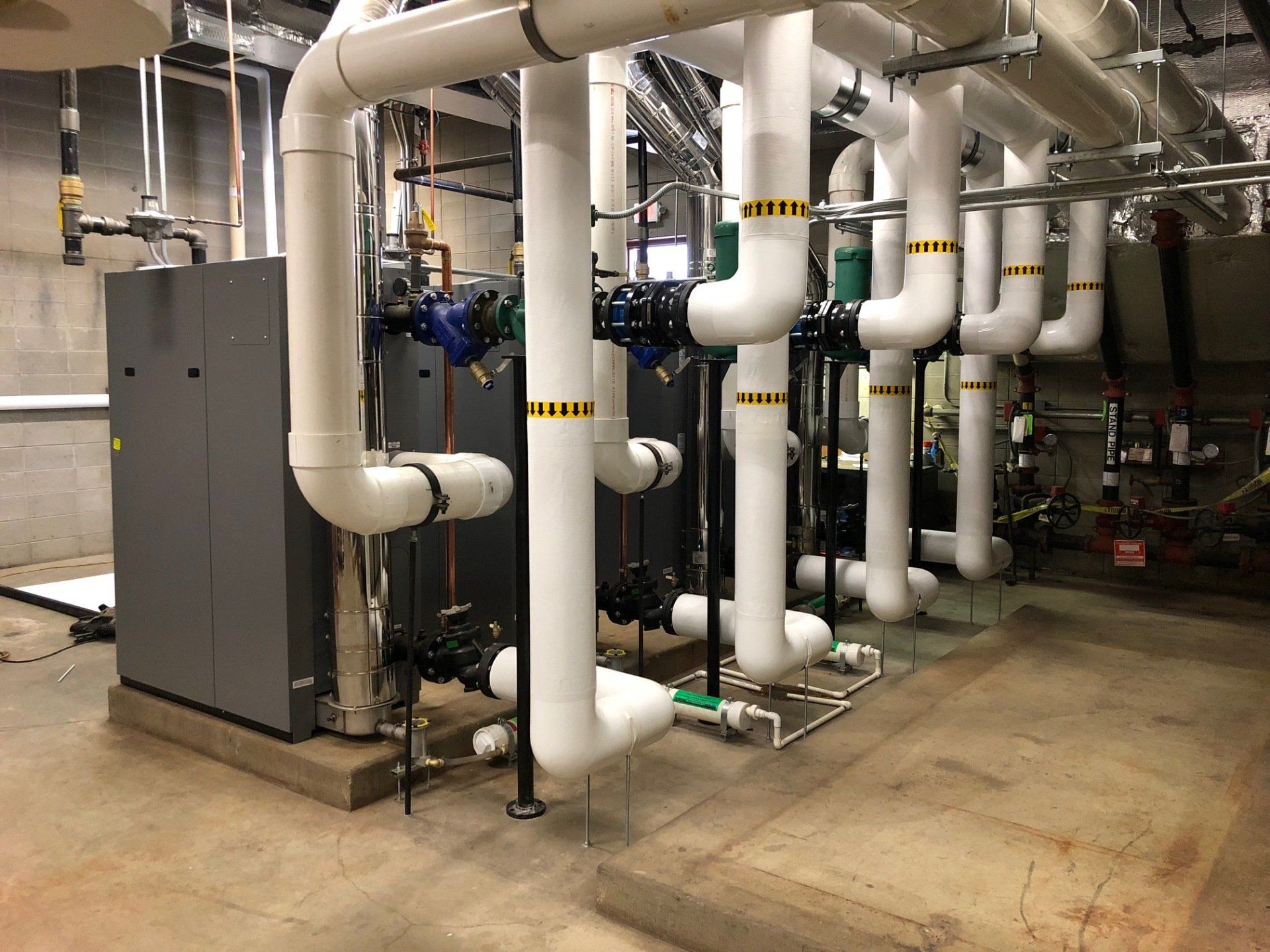 HVAC equipment in a mechanical room with white pipes, valves, and a gray cabinet on a concrete floor.
