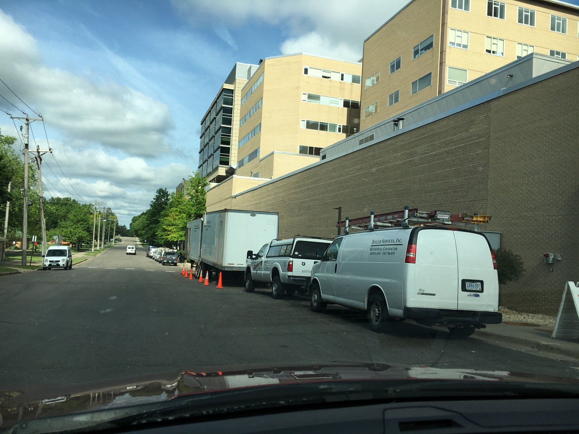 Street view with parked vehicles.  Large building in background, orange cones, and blue sky.