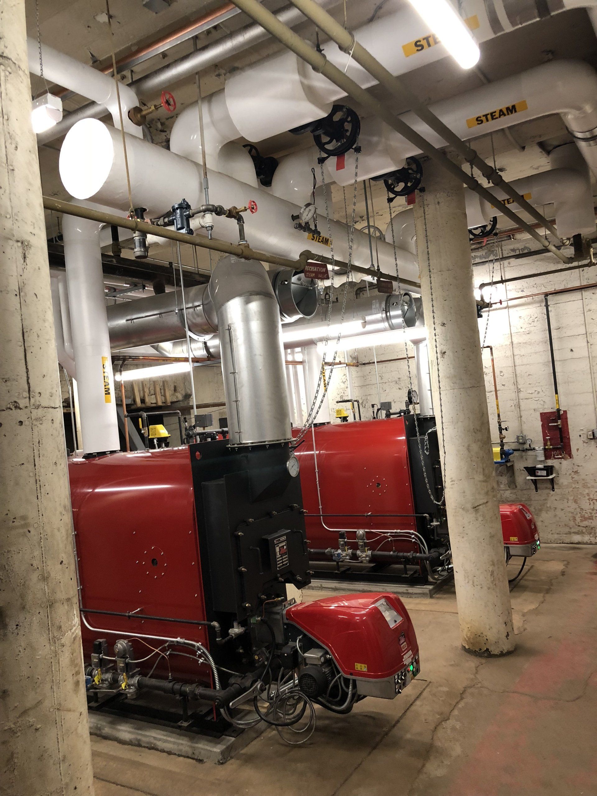 Two red industrial boilers with pipes in a basement setting.