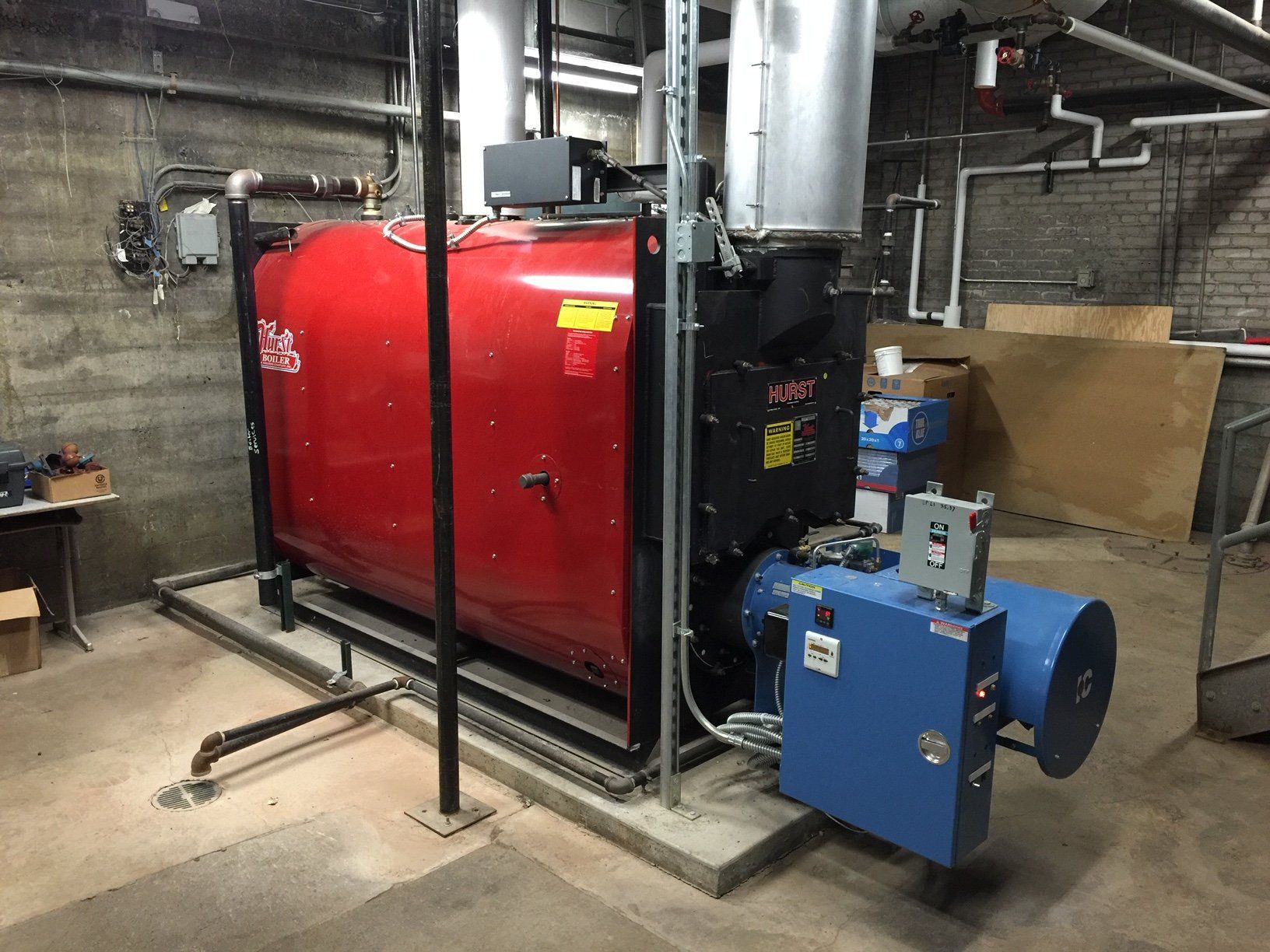 Red and black industrial boiler in a concrete room. Blue burner attached. Piping and ventilation visible.