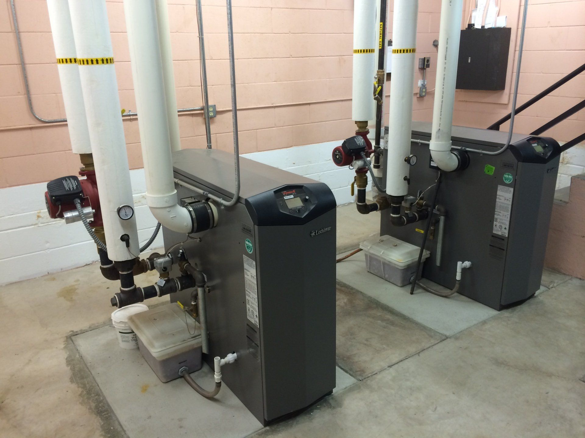 Two gray boilers with white pipes in a utility room.