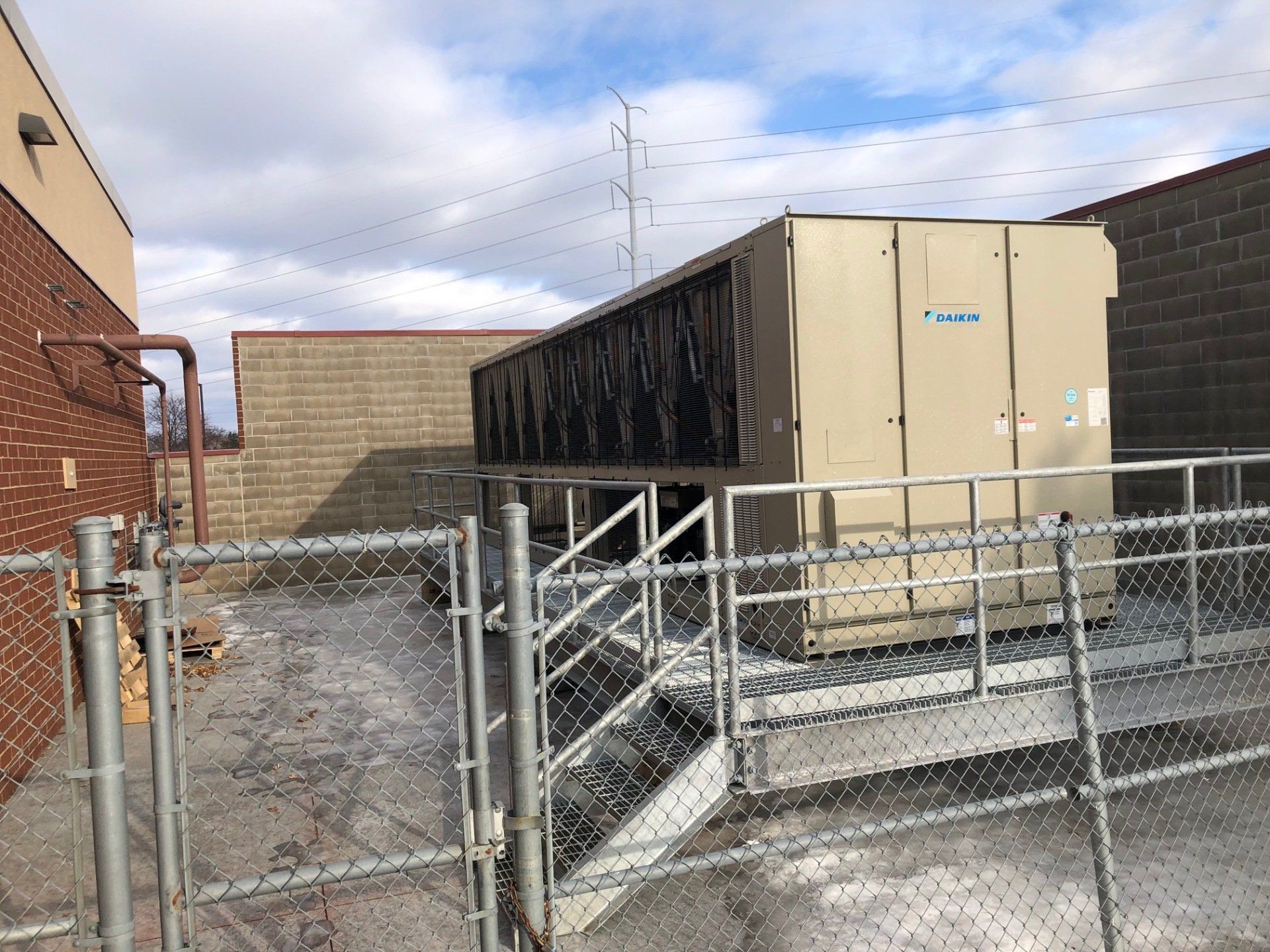 An outdoor industrial HVAC unit on a raised platform, surrounded by a chain-link fence, next to a brick building.
