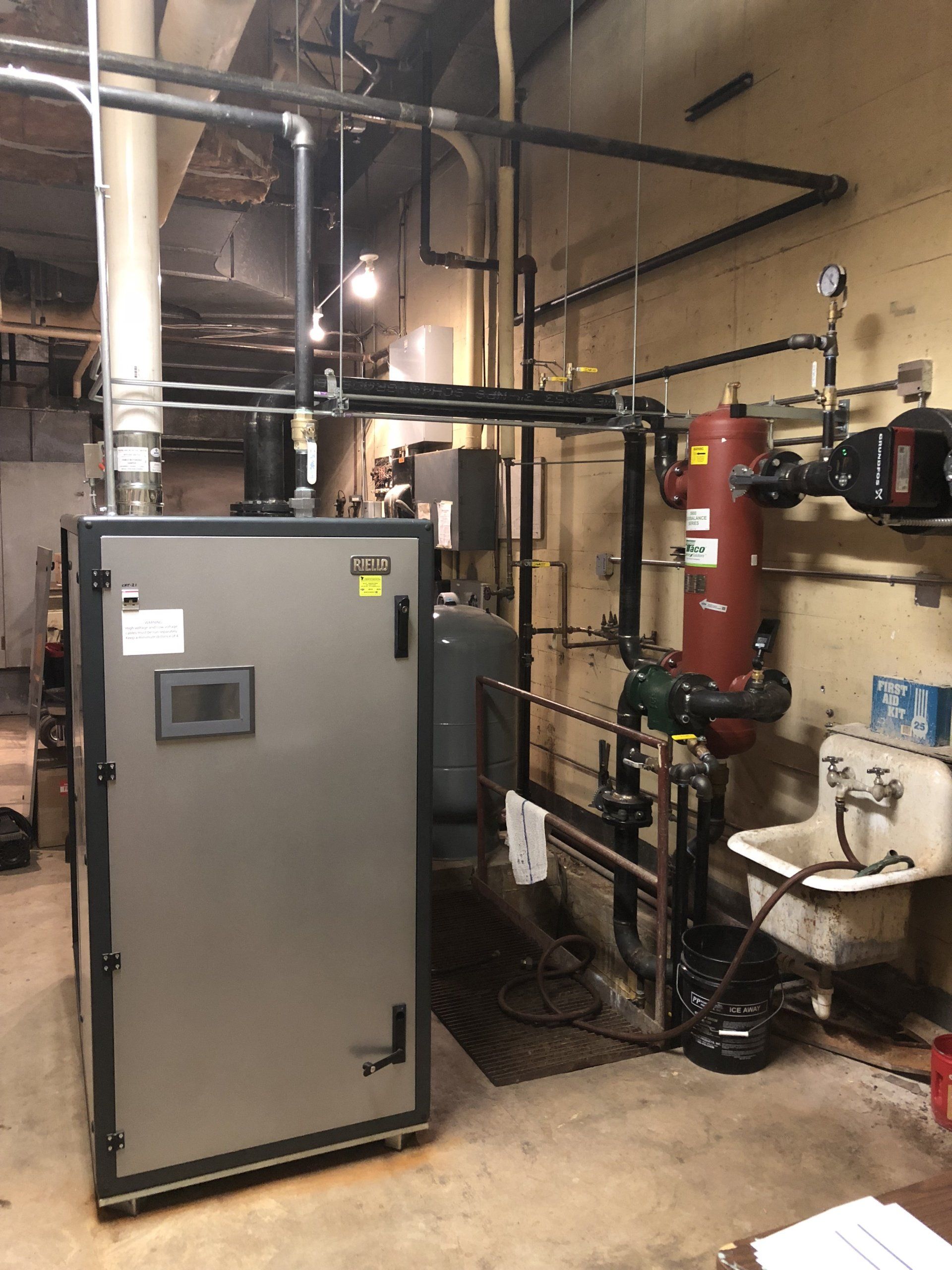 A large industrial boiler with piping in a basement setting. Grey and black tones, with a red water tank and a utility sink.