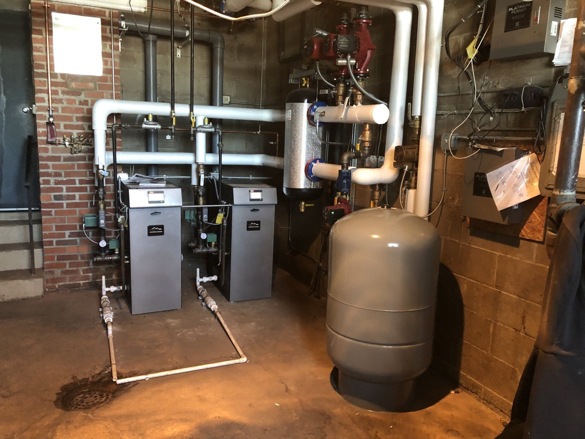 Two grey boilers with white pipes in a basement setting next to a pressure tank and brick wall.
