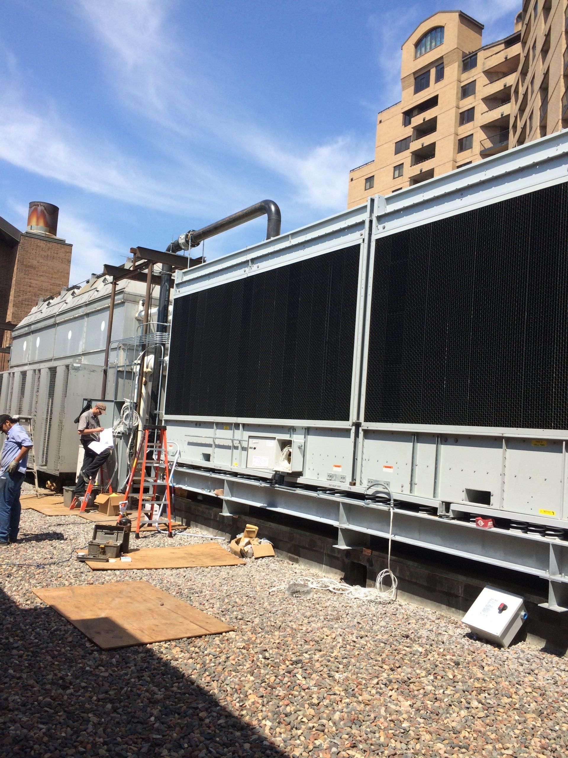 Cooling tower on a rooftop with workers, brick building in background. Sunny day.