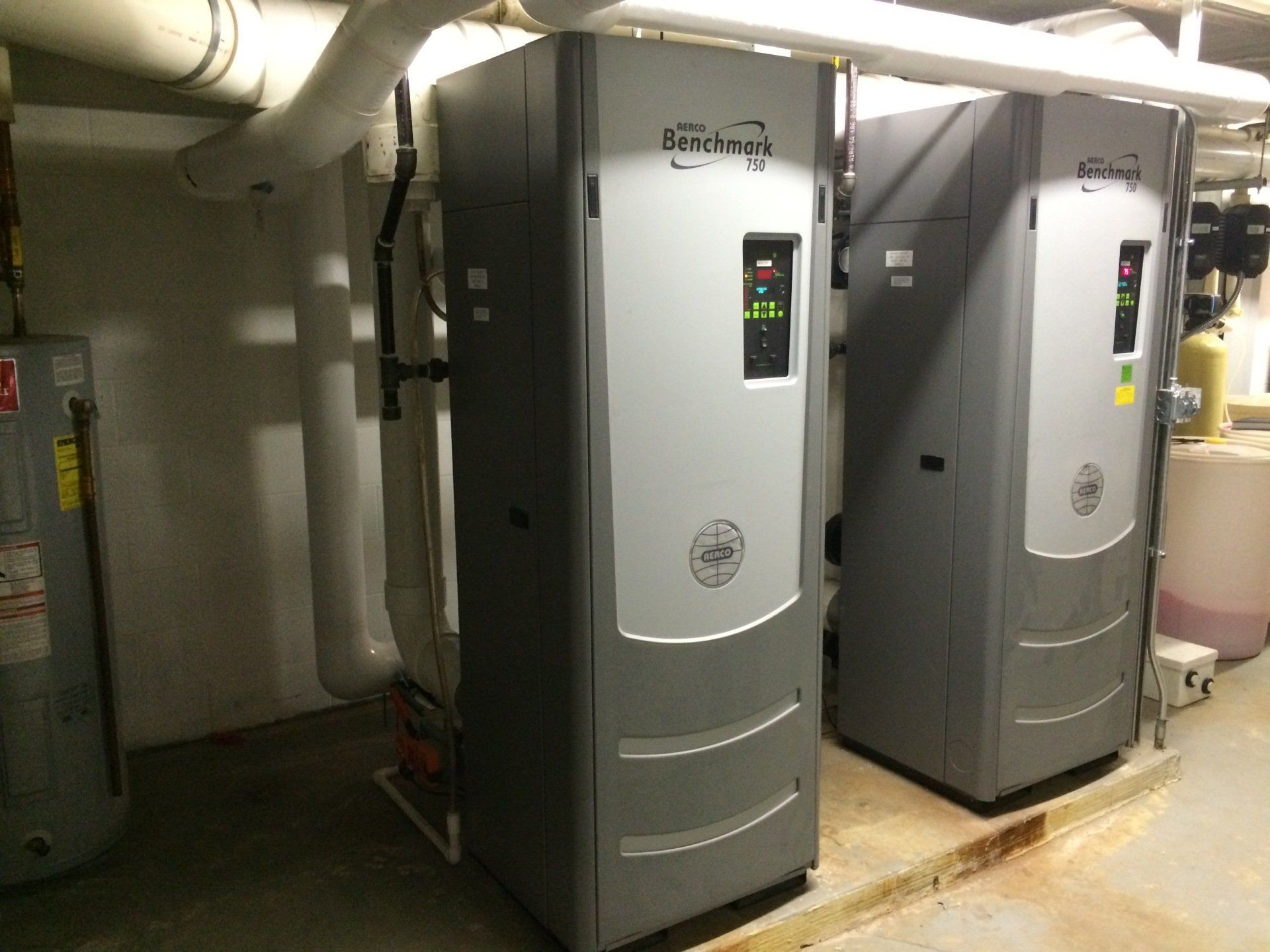 Two tall, gray industrial heating units side-by-side in a basement with pipes.