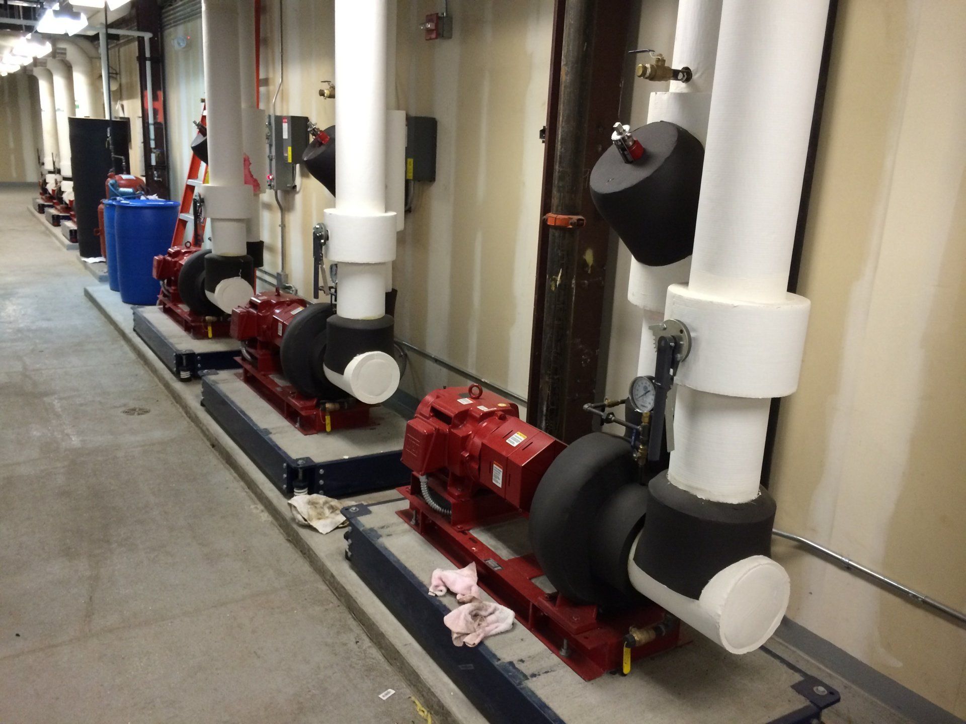 Row of red pumps with black insulation, white pipes, and blue tank in a mechanical room.
