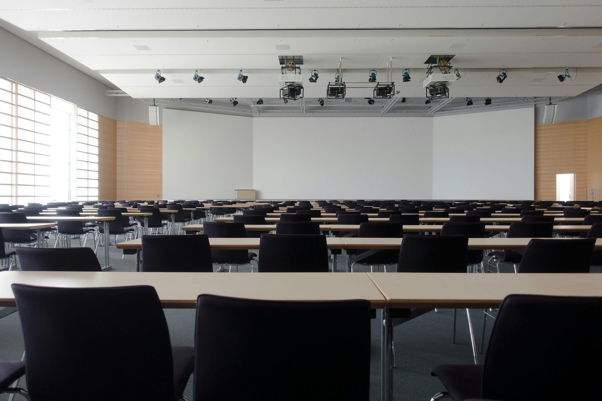 Rows of empty chairs and tables in a large conference room, with a projection screen on the wall.