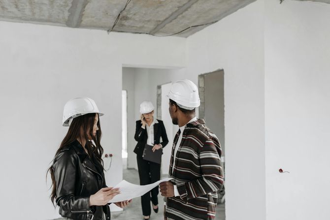 Three people wearing hard hats review blueprints in an unfinished, white-walled building. A fourth person walks in the background.