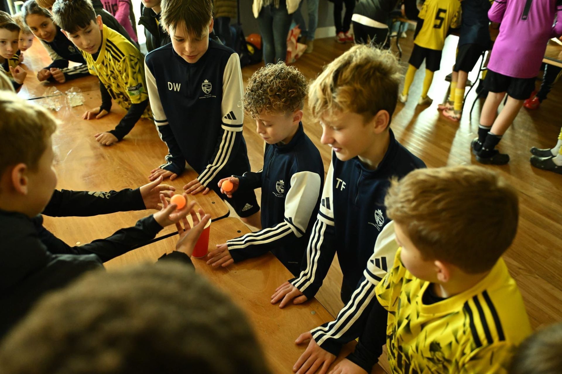 Boys in sports uniforms interacting at a table, some holding objects, indoors.