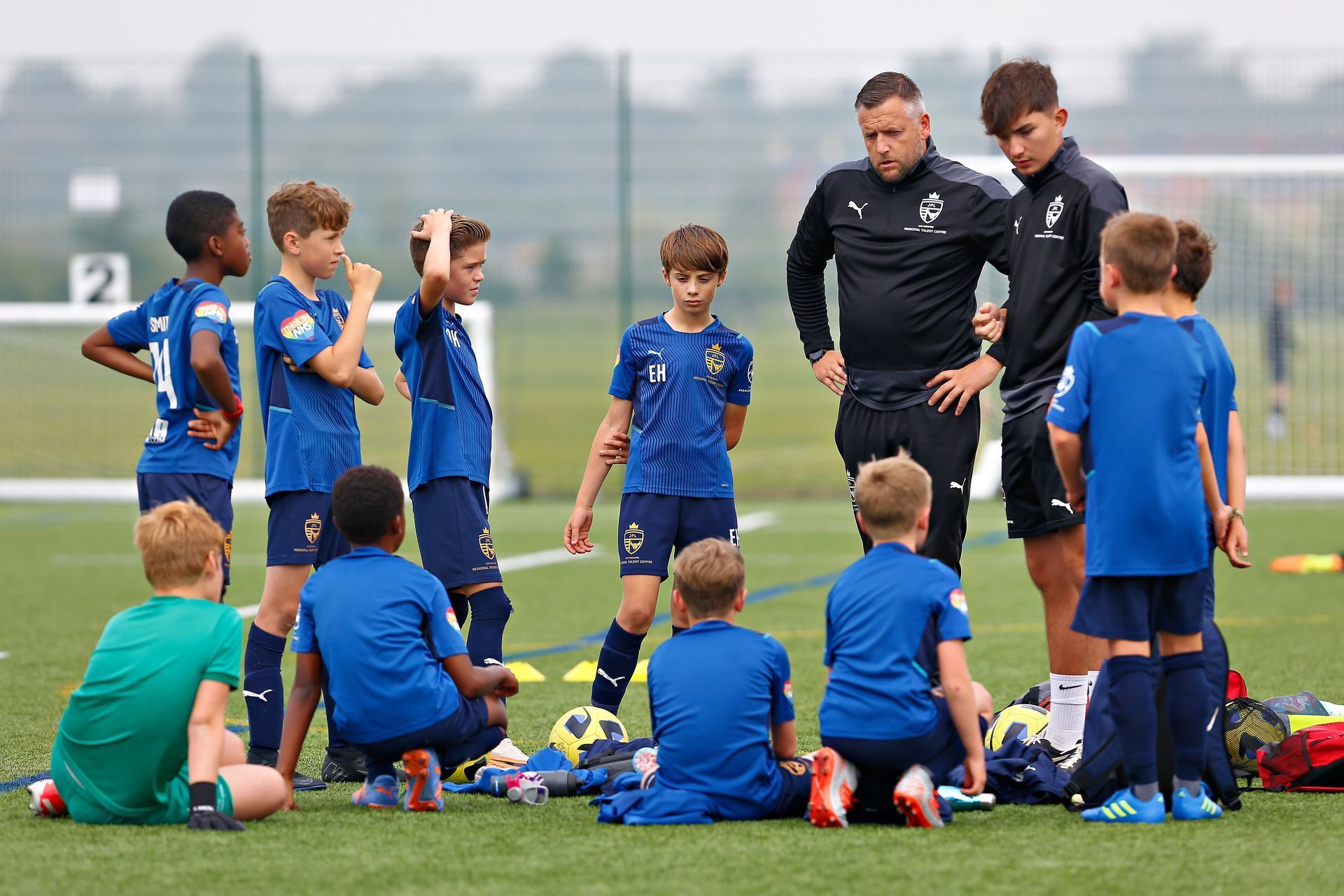 Soccer coaches instruct a youth team in blue jerseys on a green field.