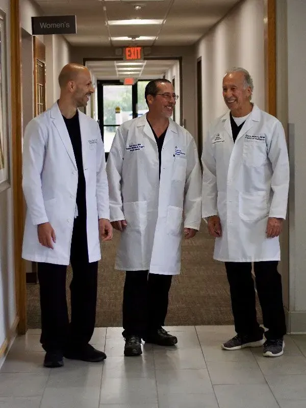 Three doctors in white coats smile in a hospital hallway.