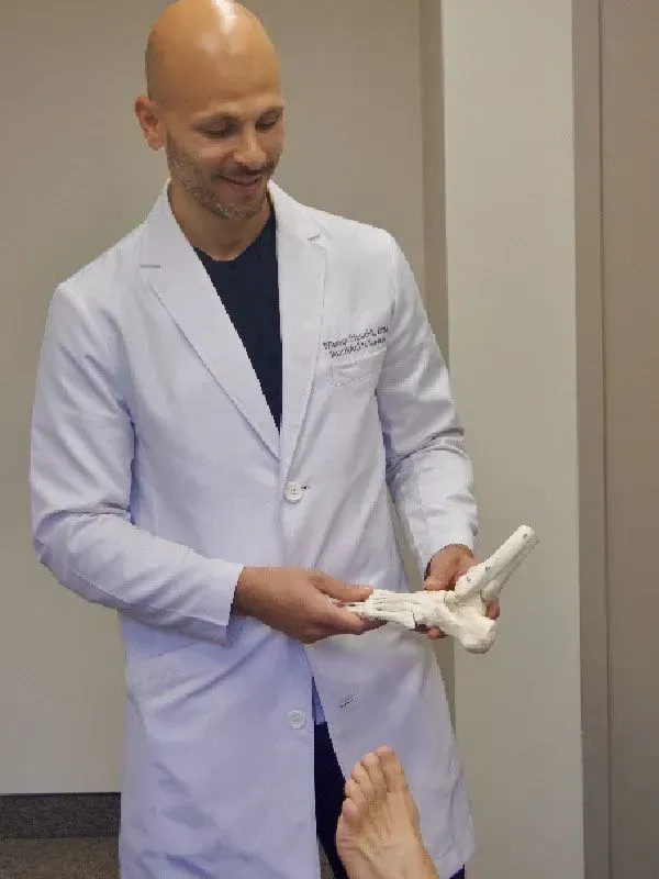Doctor in a white coat holding a foot skeleton, examining a patient’s foot. Inside a medical office.