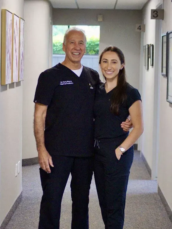 Two medical professionals in dark blue scrubs stand in a hallway, smiling, with an arm around each other.