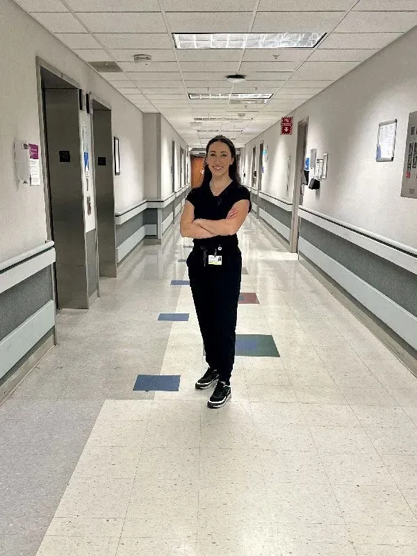 Woman in scrubs stands in hospital hallway with arms crossed.