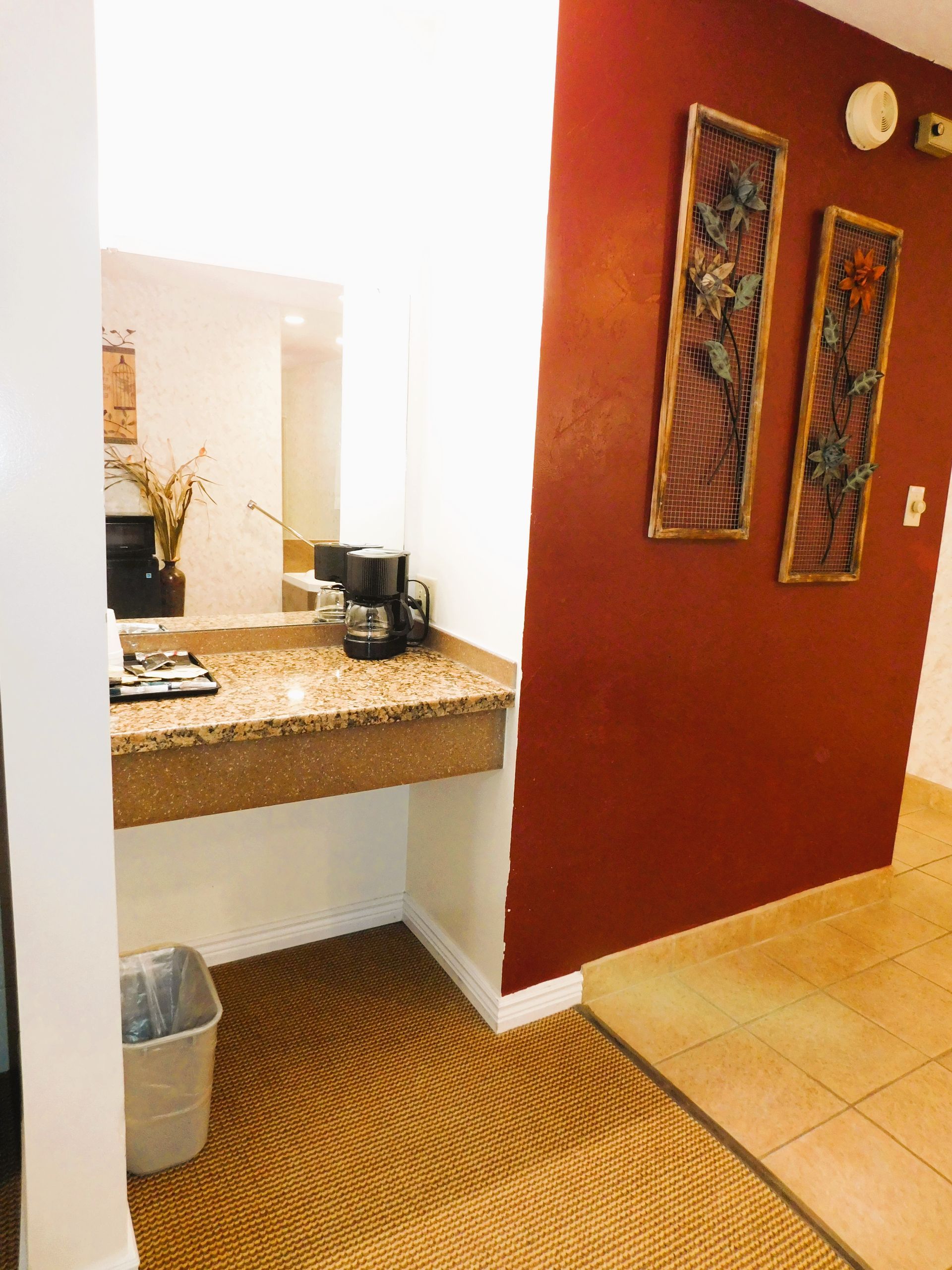 Hotel coffee station with counter, coffee maker, trash can, and red wall with framed art.